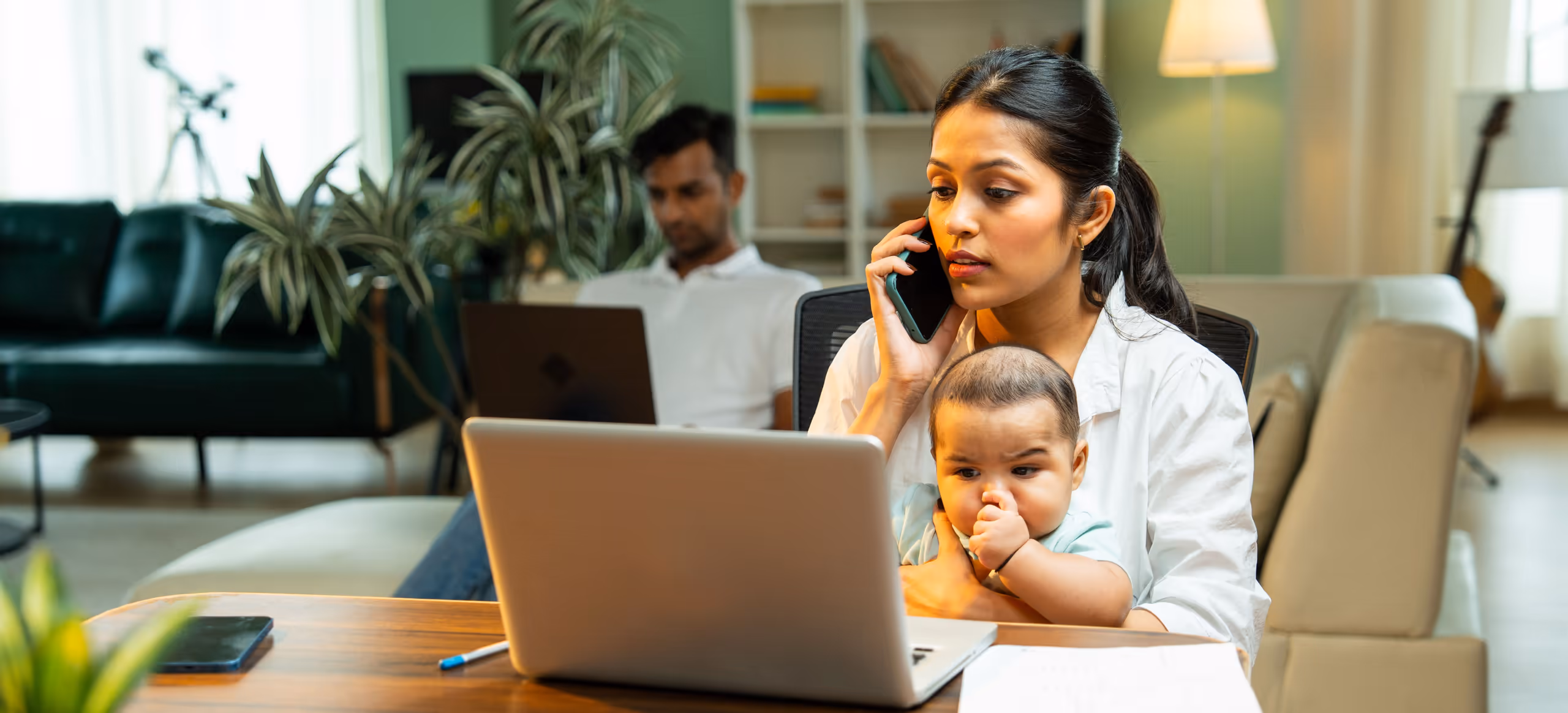 A parent on the phone at a laptop with a baby on her lap, a partner working behind her