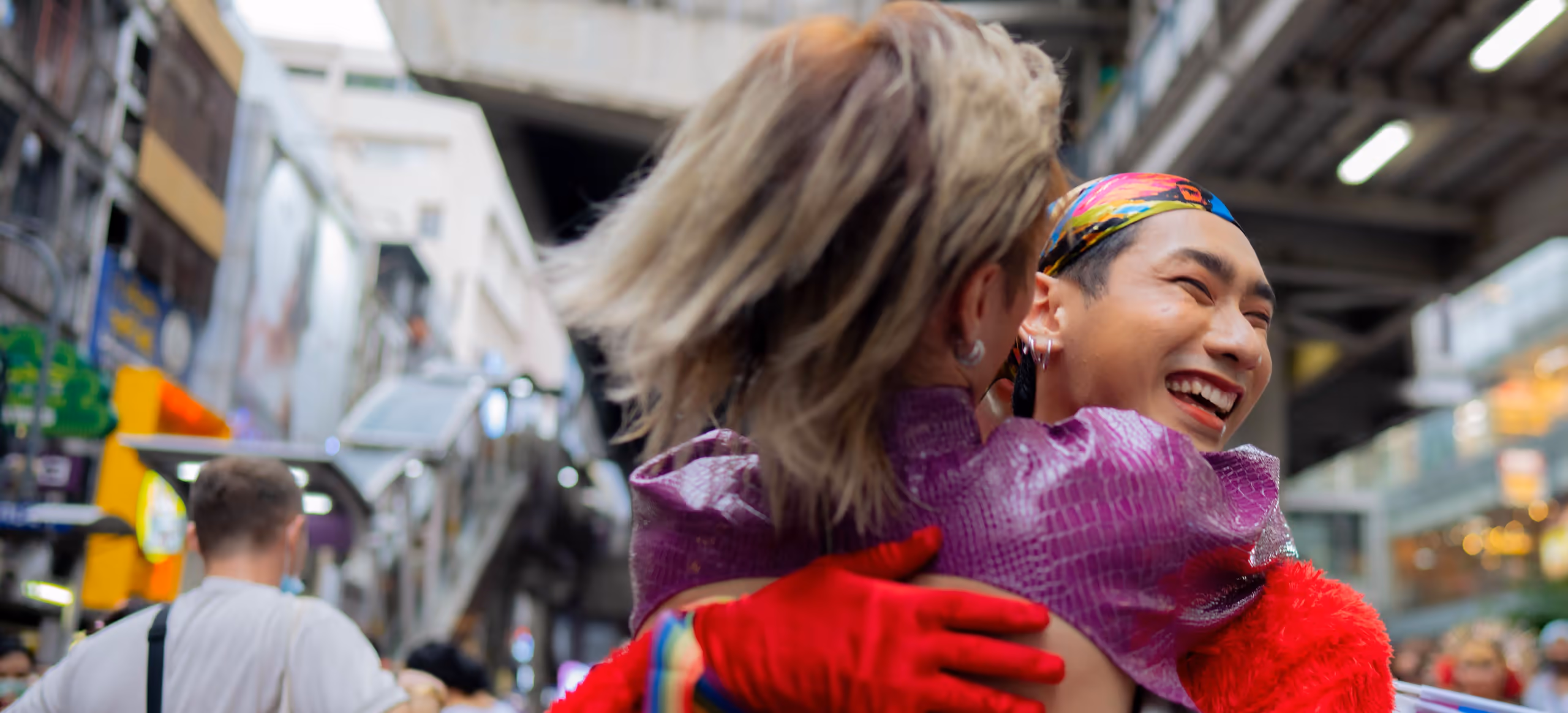 Two friends laughing as they embrace at a Pride parade, rainbow flags around them