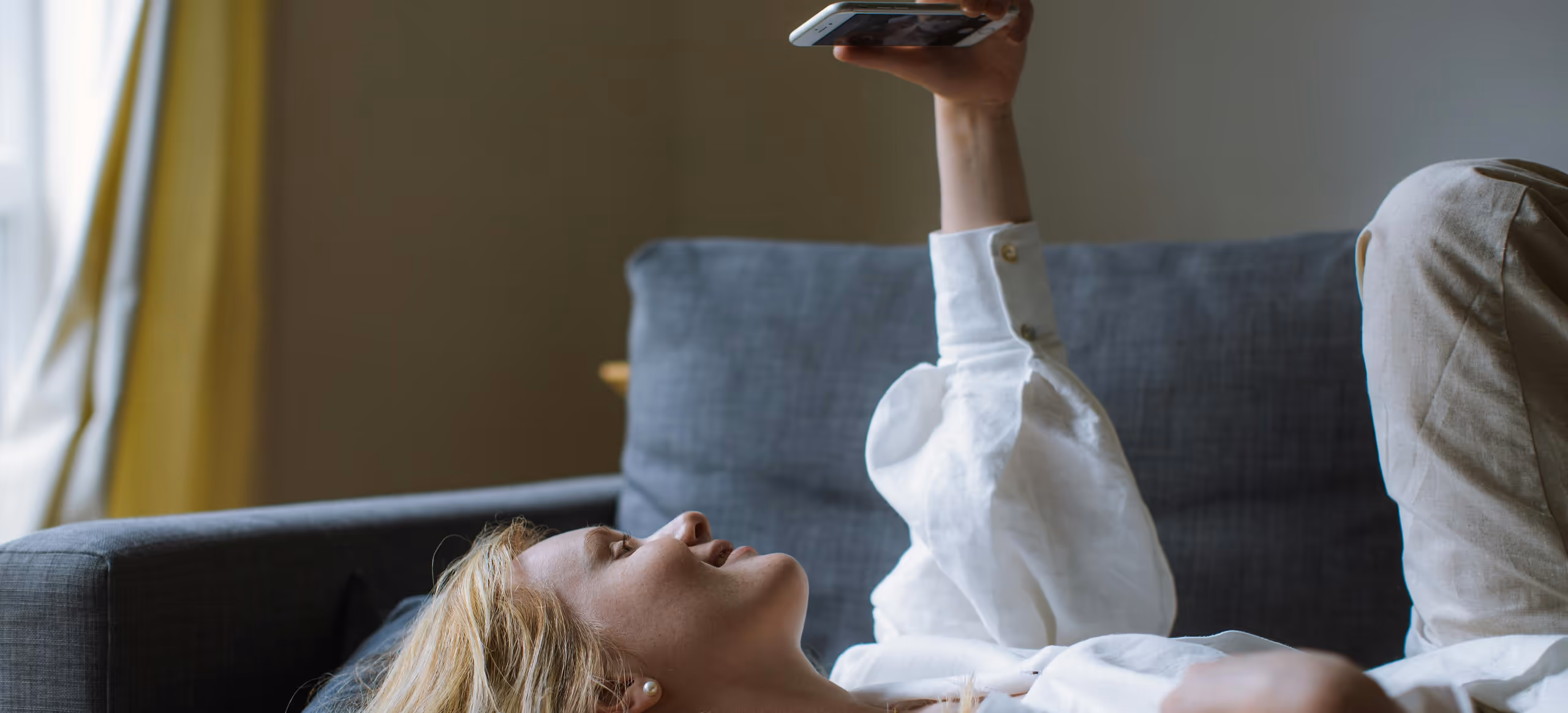 A woman lying back across a sofa, holding a phone above her at arm’s length