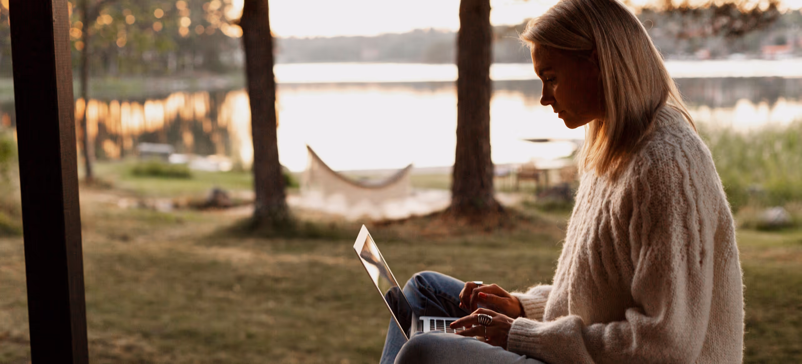 A woman sitting cross-legged on a porch with a laptop, a lake behind her at sunset