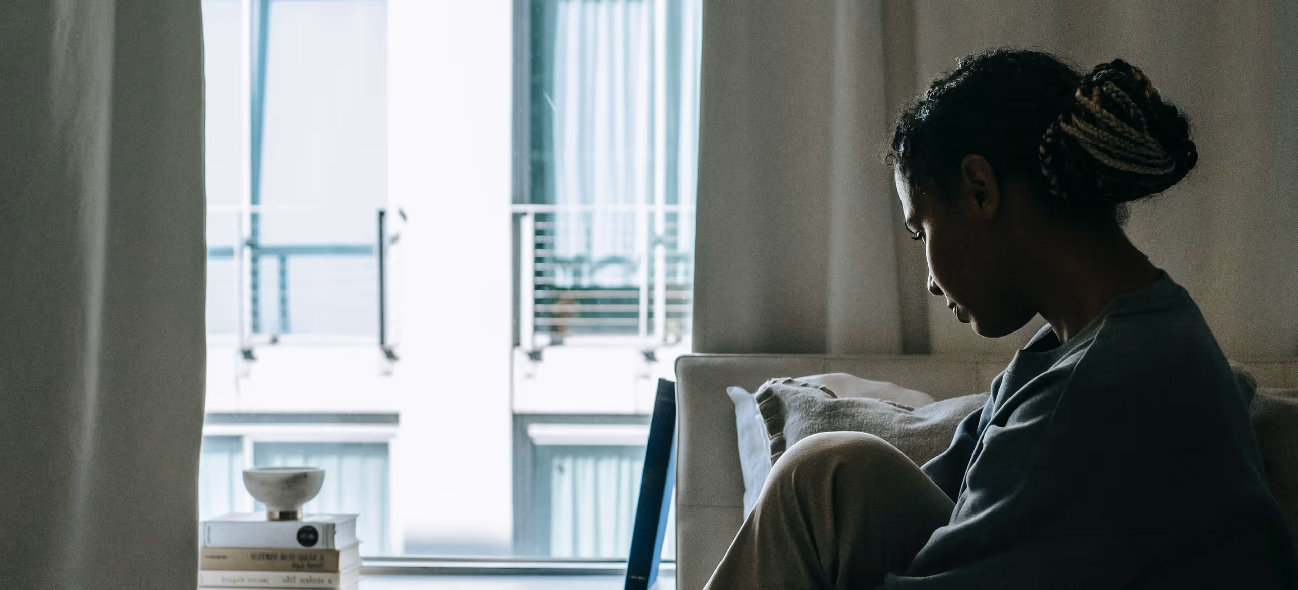 A woman sitting on a bed with her knees drawn up, looking down, daylight from a window beside her